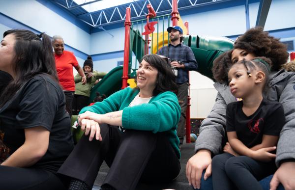 Woman sits on floor, smiling, next to families.