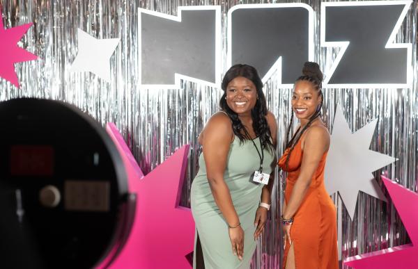 Two women in formal dresses smile for the camera in front of a large block lettering reading "NAZ"