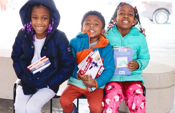 Three young children sit in their winter jackets holding books