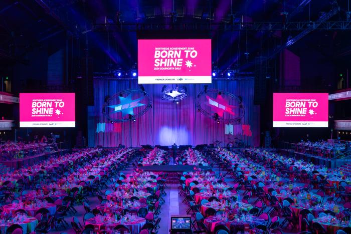 A large room full of tables, lit purple and pink, with three different screens with "Born to Shine" on them
