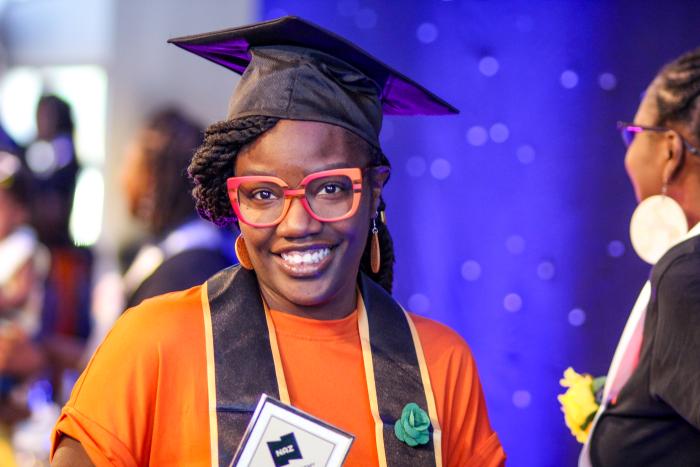 Woman in glasses, holding award, wearing graduation stole and cap smiling at camera.