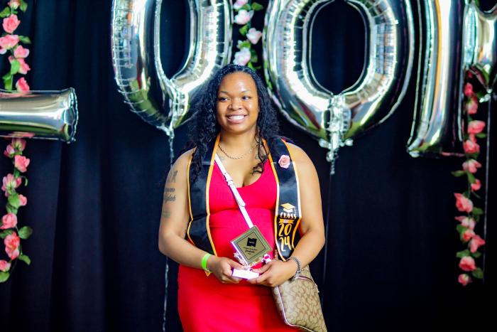 Woman stands in front of balloons with award wearing graduation stole.