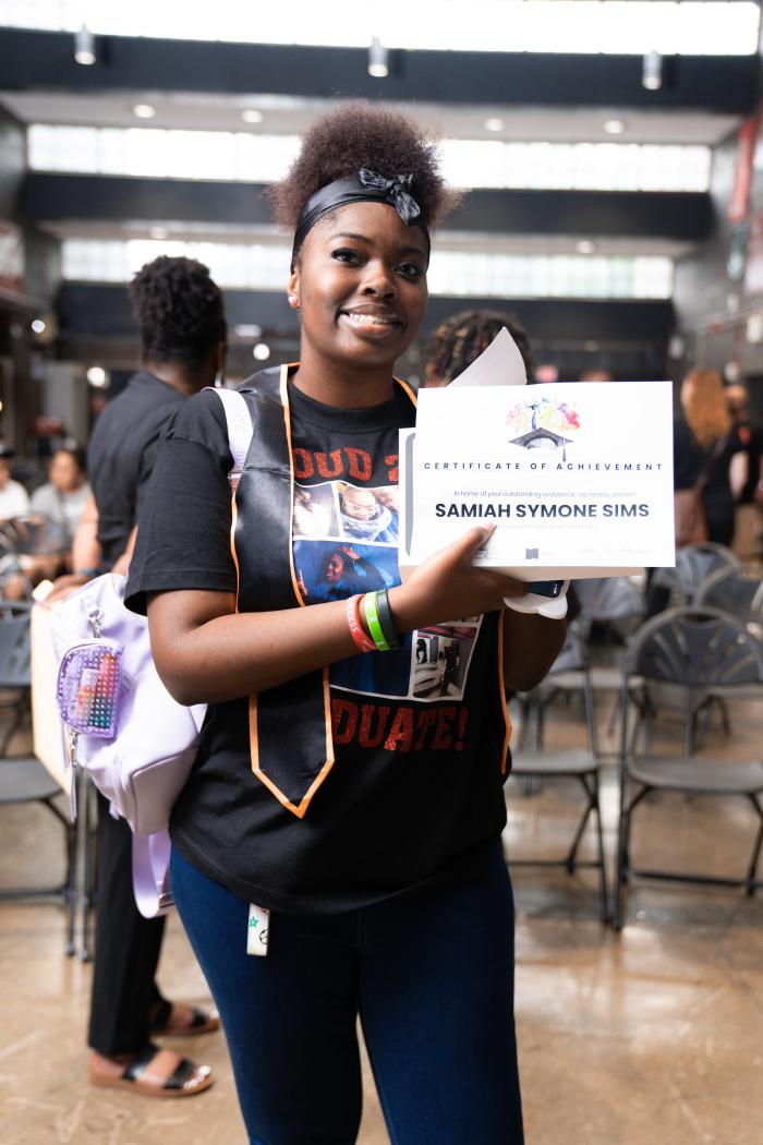 A scholar holds her certificate while smiling, wearing a stole.