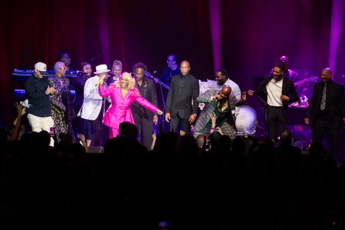 Stage full of people in fancy dress dancing with a woman in a pink suit singing into a microphone.