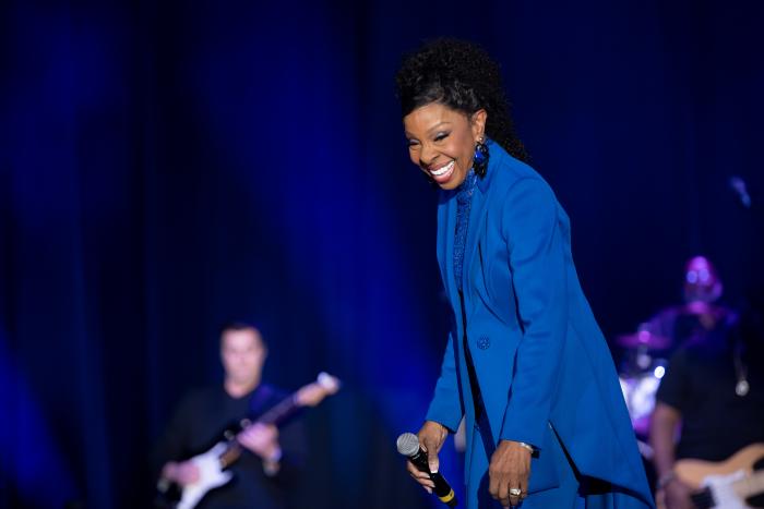 Woman in blue suit, holding microphone, smiling with musicians in the background.