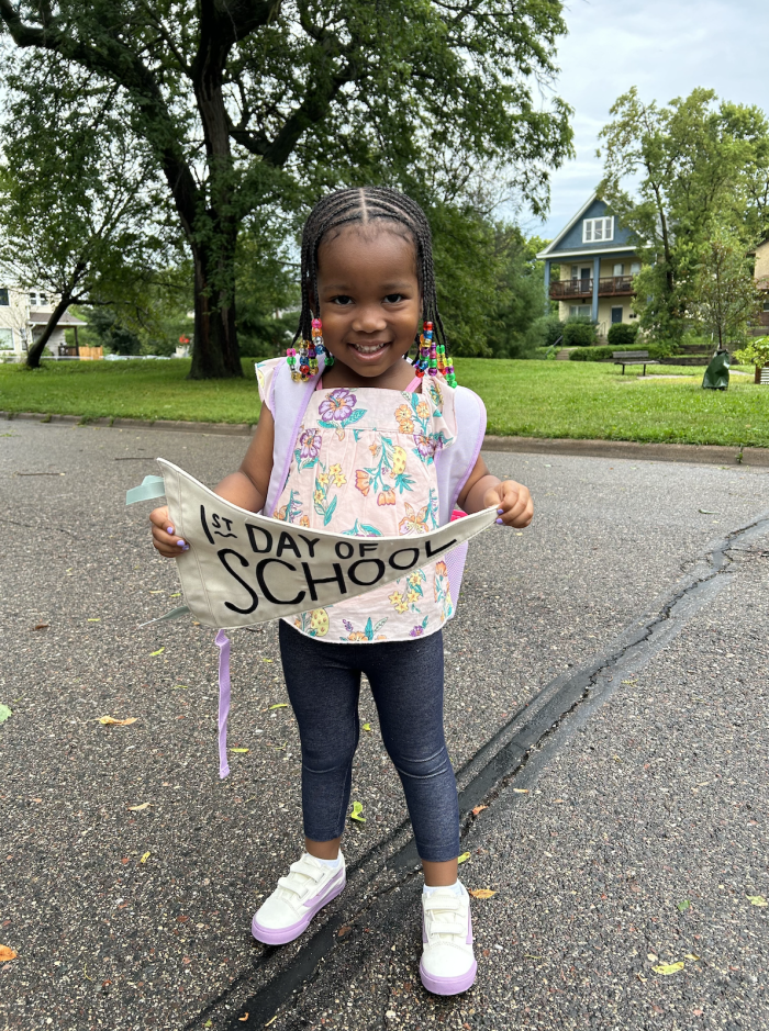 Young girl stands with a backpack on and "First Day of School" banner in-hand