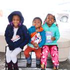 Three young children sit in their winter jackets holding books